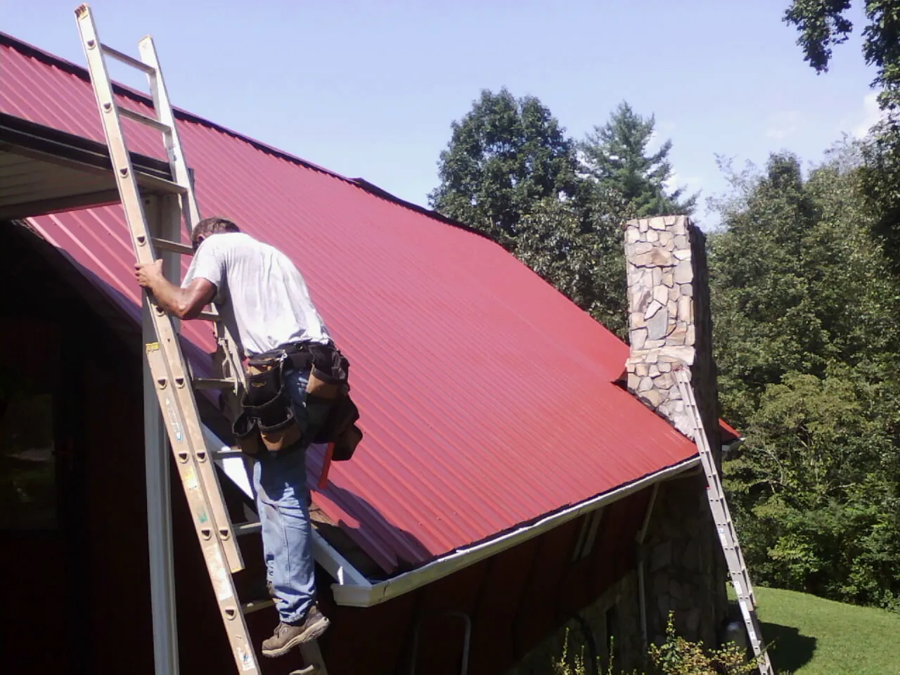 Roofer on a ladder beside a red metal roof and stone chimney — Soffit & Fascia Repair by Roofing Brothers in Doylesburg